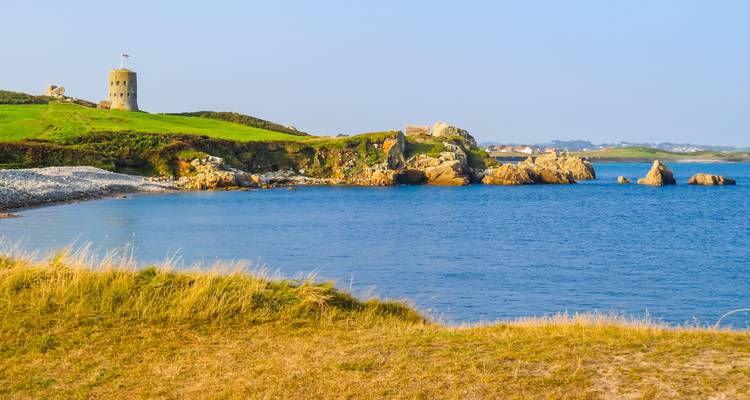 Stone tower and rocky coastline meet calm blue sea under clear morning light on Guernsey.