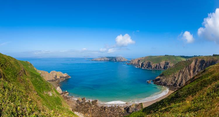 Panoramic view of a secluded crescent beach surrounded by steep green cliffs and turquoise water.