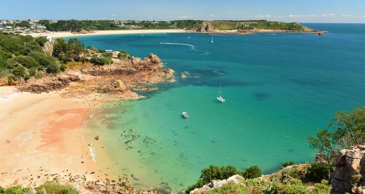 Turquoise bay with sandy beach and sailboats framed by green cliffs viewed from above.
