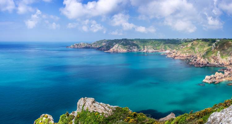 Wide coastal panorama of rugged green headlands and vivid blue sea under scattered clouds.