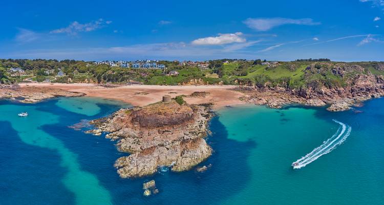 Aerial shot of a rocky promontory, golden beach and speedboat carving a white wake in turquoise water.