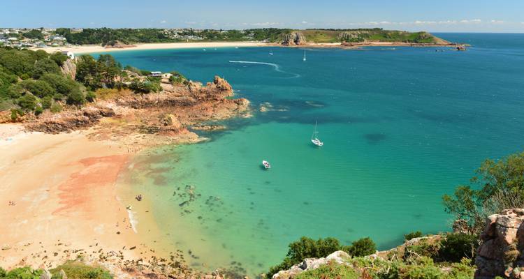 Clear view from clifftop over sandy cove with emerald water, dotted boats and surrounding greenery.