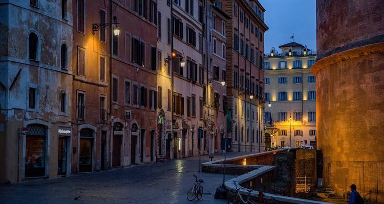 Empty street with historical buildings lit by street lamps during evening.