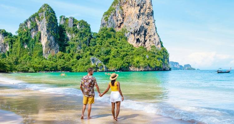 Pareja tomada de las manos en una playa tropical con acantilados.