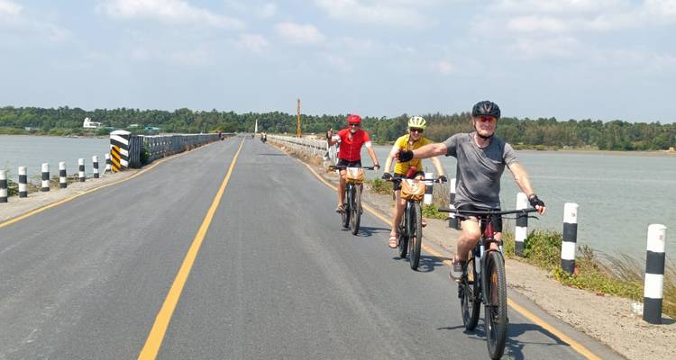 Drie fietsers op een weg met een waterlichaam en een bosgebied.