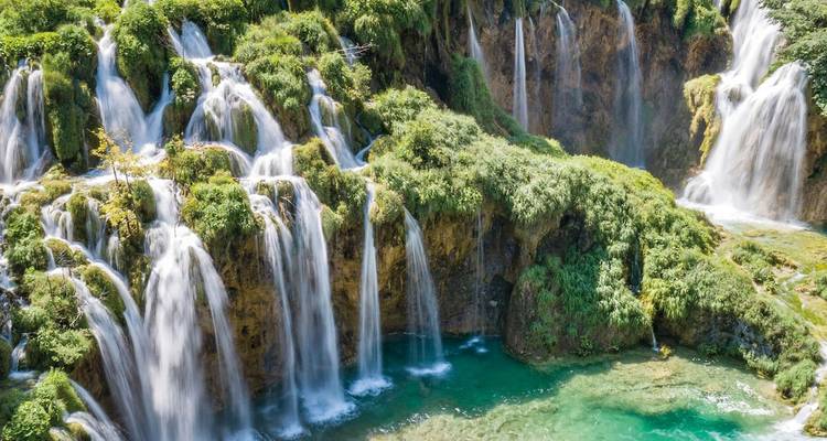 Repetition: vibrant aerial of turquoise pools and gushing falls amid dense forest at Plitvice Lakes.