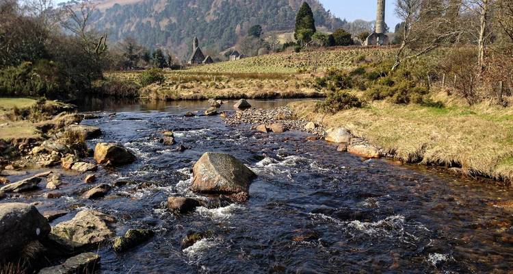 Shallow rocky stream flowing through a grassy valley with an ancient round tower and stone ruins in the background