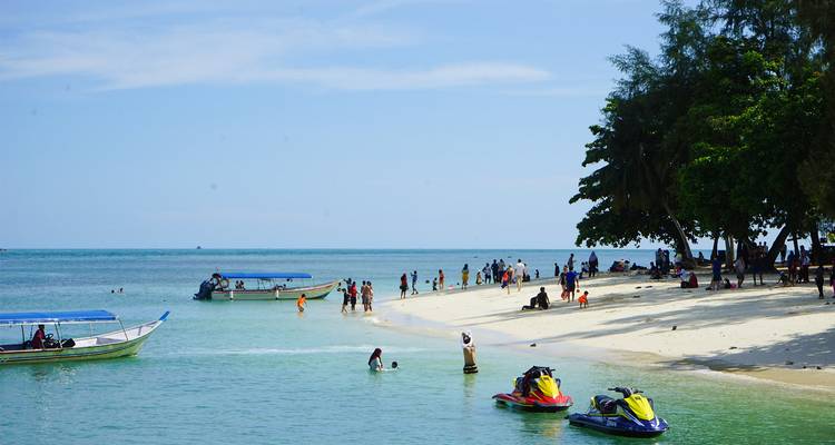 Los visitantes de la playa nadan y juegan cerca de botes y motos acuáticas en una orilla tropical de arena blanca.