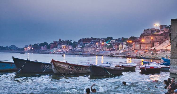 Boote auf dem Ganges mit den beleuchteten Ghats von Varanasi im Hintergrund in der Dämmerung.