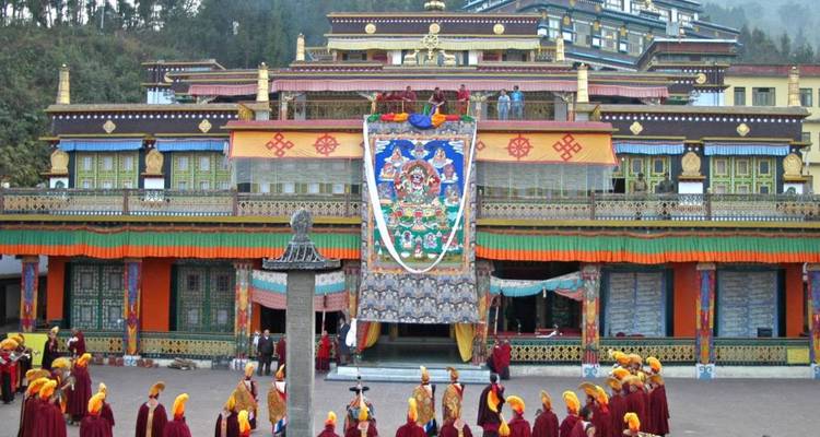 Des rangées de moines bouddhistes vêtus de robes écarlates se rassemblent devant un monastère multiétage aux couleurs vives affichant une grande bannière thangka verticale.