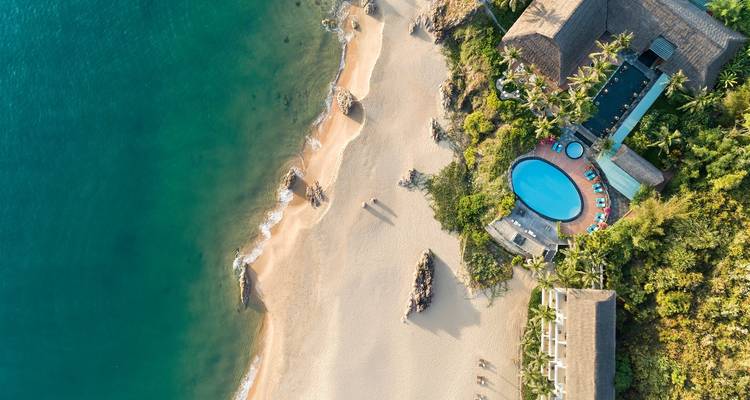 Luchtfoto van een badplaatsresort aan het strand met zwembad en omringende groenvoorzieningen.