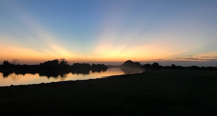 Zonnestralenklinken zich uit over een stille rivier met donkere boomsilhouetten aan de oever.