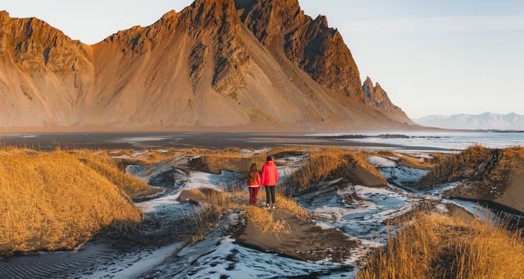 Una pareja caminando en una playa de arena con picos montañosos puntiagudos al fondo.