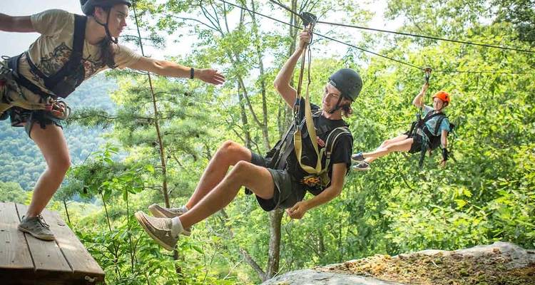 Drei Personen auf einer Seilrutsche-Plattform in einem bewaldeten Gebiet.