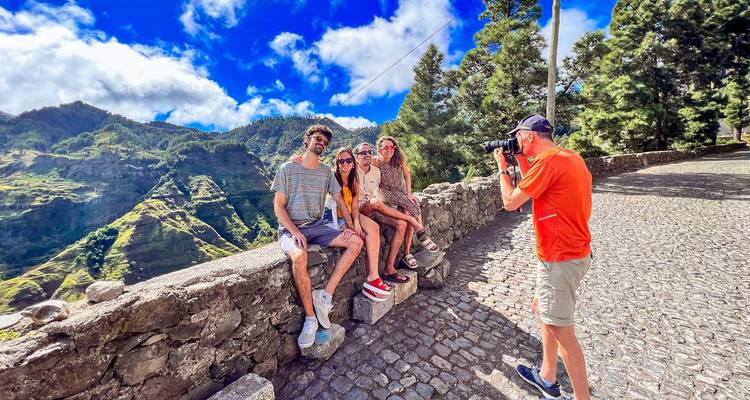 Grupo de amigos sentados en una pared de piedra mirando hacia unos paisajes montañosos espectaculares mientras una persona los fotografía