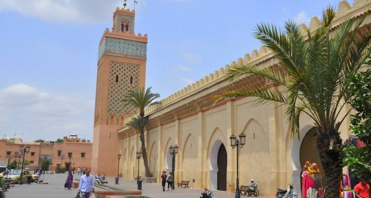 Tagesansicht der Koutoubia-Moschee neben einer langen ockerfarbenen Mauer, die von Palmen und Fußgängern gesäumt ist.