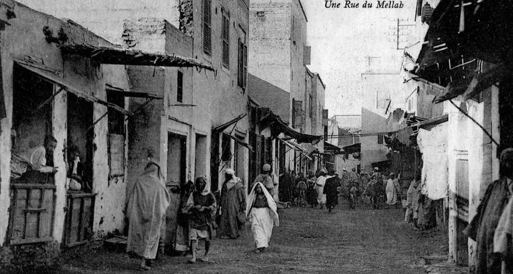 Historische Schwarz-Weiß-Fotografie einer belebten Mellah-Straße, gesäumt von bröckelnden Gebäuden und Einheimischen in traditioneller Kleidung.