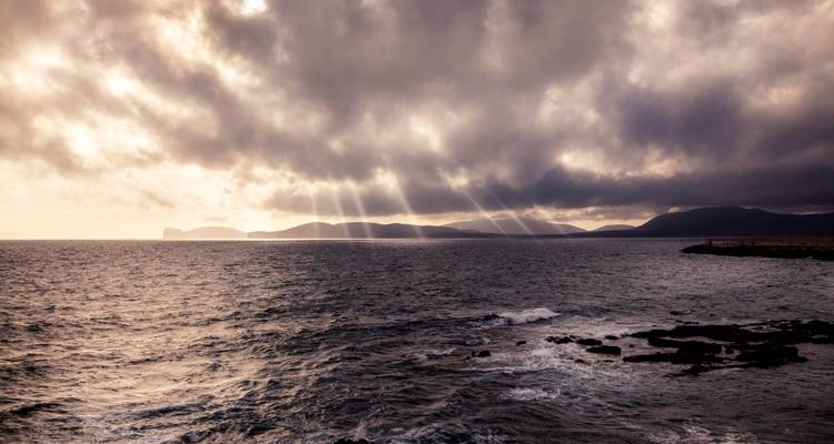 Paysage marin aux teintes sombres avec des rayons de soleil perçant les nuages épais au-dessus d'une côte accidentée et de eaux agitées.