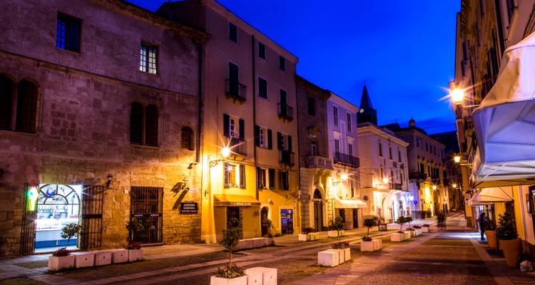 Rue historique tranquille à Alghero illuminée par les lumières chaudes du soir sous un ciel crépusculaire bleu profond.