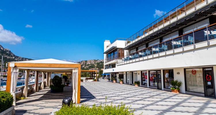Promenade et place waterfront modernes avec boutiques et marina adossées aux collines rocheuses à Porto Cervo.