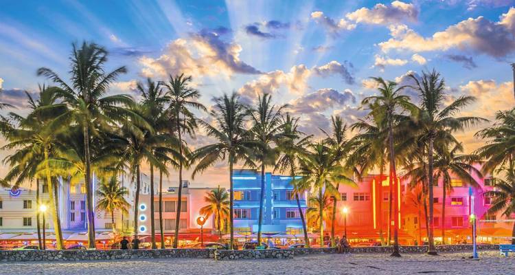 Edificios de neón en tonos pastel y palmeras que se mecen brillan al atardecer en la animada Ocean Drive con un cielo colorido arriba.