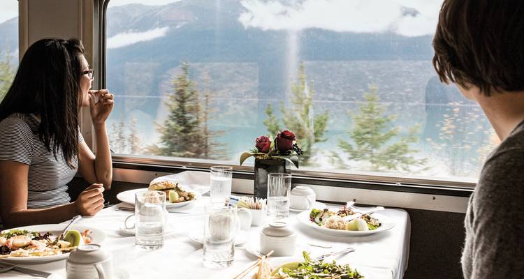 Passagiere genießen eine Mahlzeit mit malerischem Bergblick aus einem Zugfenster.