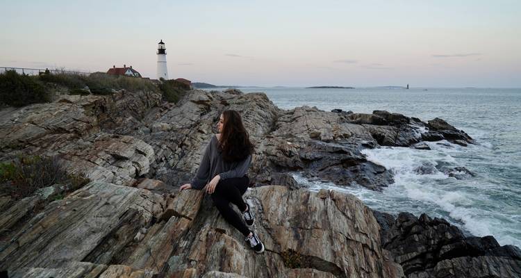 Person, die auf Felsen am Meer mit einem Leuchtturm sitzt.