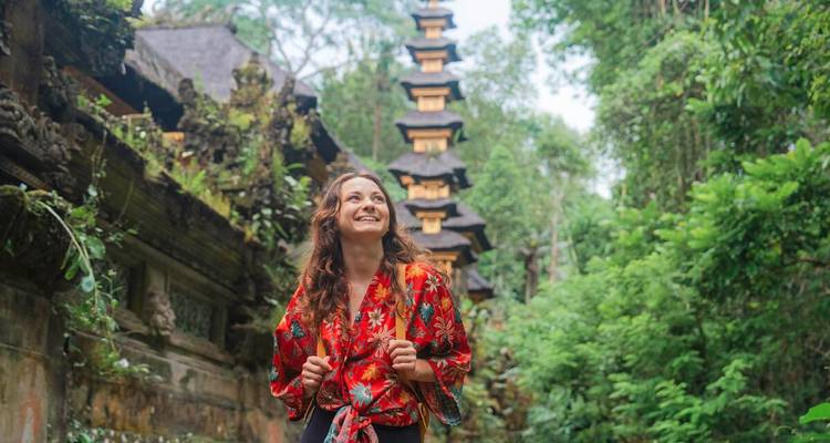 Junge Frau in rotem Kimono-Top mit Blumenmuster lächelnd in einem üppigen balinesischen Dschungeltempel.