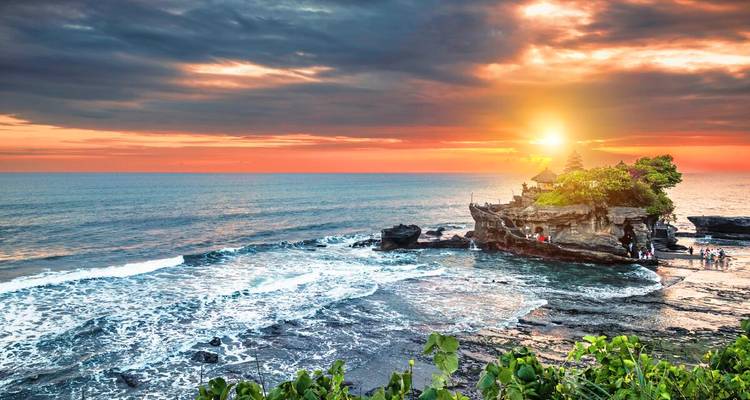 Die Sonne geht hinter dem Tempel Tanah Lot unter, der auf einem felsigen Vorsprung im Meer thront, während Wellen unten aufschlagen.