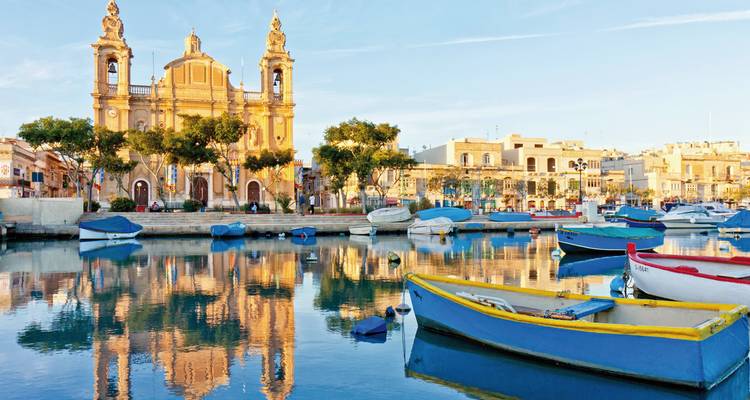 A charming waterfront with small boats and an ornate church building reflecting on the water.