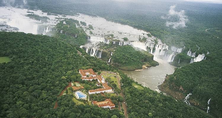 Luftaufnahme eines großen Hotelkomplexes neben blühenden Wäldern und Wasserfällen.