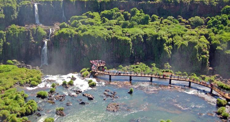 Menschen gehen auf einer Brücke über einen Fluss in der Nähe eines bewaldeten Gebiets mit Wasserfällen.