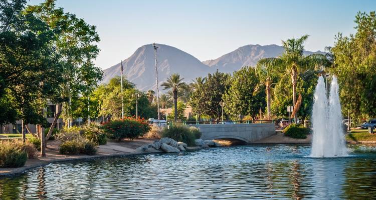 Parque pintoresco con un estanque, fuente y montañas al fondo.