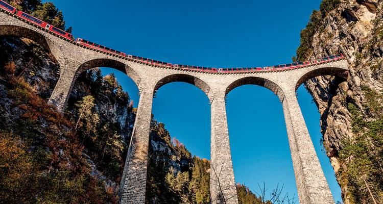 Train crossing a tall viaduct amidst towering cliffs and blue sky.