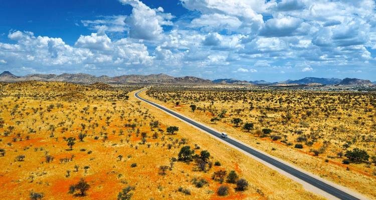 Eine lange, gerade Autobahn durchschneidet Namibias ausgedehnte rote und gelbe Wüstenebenen in Richtung entfernter Hügel.