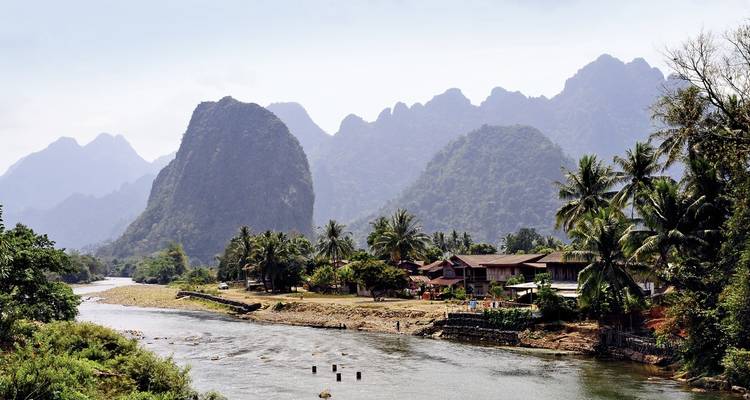 Scène de village au bord d'une rivière avec des montagnes verdoyantes.