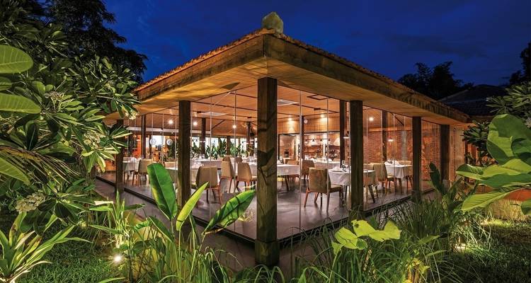 Elegant glass-walled restaurant lit up at night with surrounding greenery.
