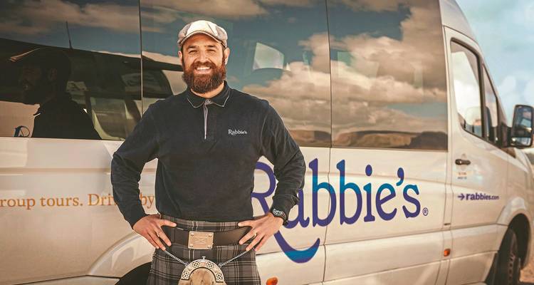 Hombre con falda escocesa parado frente a un autobús con un logo, sonriendo a la cámara.