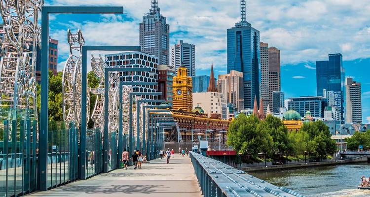 Fußgänger überqueren eine moderne Fußgängerbrücke, während sich Melbournes Skyline mit dem Uhrturm der Flinders Street Station und gläsernen Wolkenkratzern dahinter erhebt.