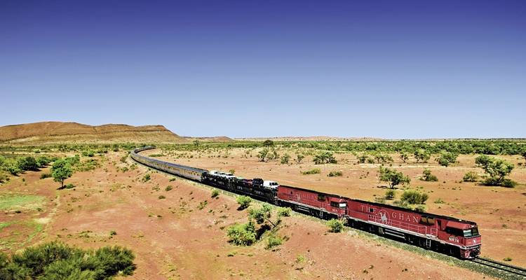 Die Ghan-Lokomotive führt einen langen Zug durch eine Wüste mit roter Erde, die mit niedrigen Sträuchern bewachsen ist, unter einem tiefblauen Himmel.