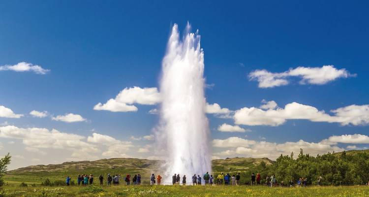 Colonne de geyser imposante jaillissant vers le ciel avec des touristes l'observant sur un fond de ciel bleu profond et de collines ondulantes.