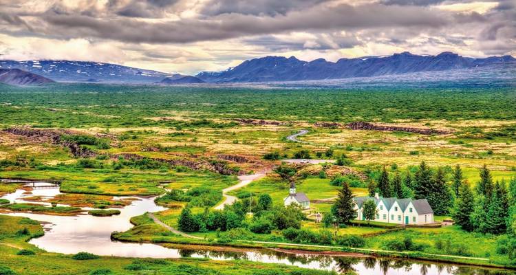 Uitgestrekte groene vallei doorspekt met rivieren en een kleine witte kerk onder dramatische luchten in het Þingvellir gebied.