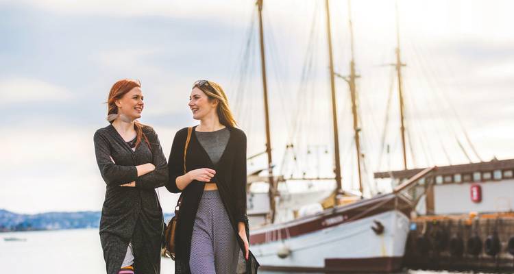 Zwei Personen, die entlang einer Uferpromenade gehen, mit einem Schiff im Hintergrund.