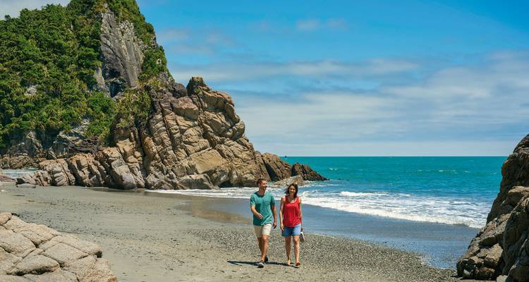 People walking along a beach with rocky cliffs and clear blue skies.