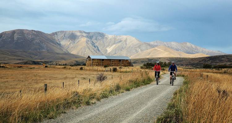 Cyclists riding on a path with mountains and a barn in the background.