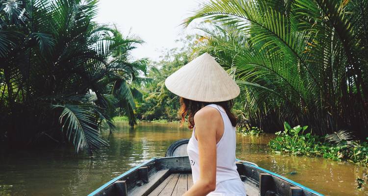 Person wearing a conical hat in a boat navigating a narrow river.