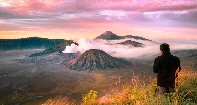 Person, die bei Sonnenaufgang auf die rauchenden Vulkane des Mount Bromo blickt.