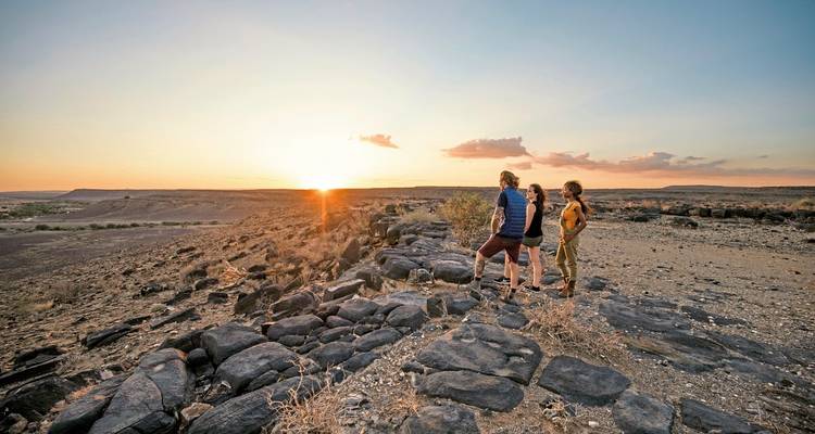 Drei Wanderer stehen auf schwarzen Vulkangesteinen und beobachten den Sonnenuntergang über einem weiten, trockenen Tal.