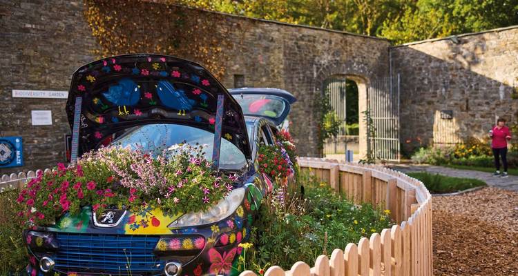 A decorated car with flowers in a garden, a person walking in the background.
