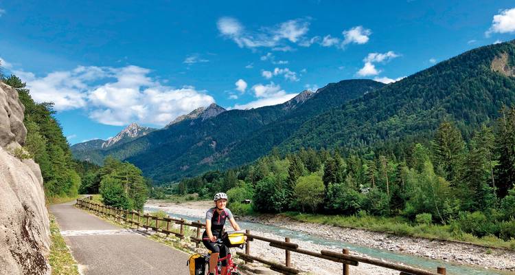 Bicyclist on a path along a river with mountains in the background.